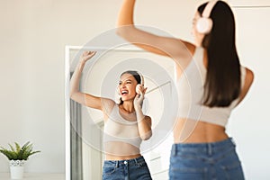 Playful young lady wearing headphones singing and dancing, listening her favourite music near mirror