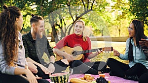 Playful young people are singing and moving hands when beautiful girl is playing the guitar during picnic in park on