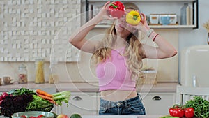 Playful young woman smiling covering eyes by yellow and red pepper in the home kitchen. Healthy lifestyle and dieting