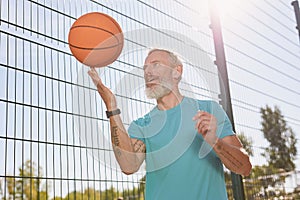 Playing basketball outdoors. Happy elderly man in sportswear spinning a basketball ball on his finger while standing at