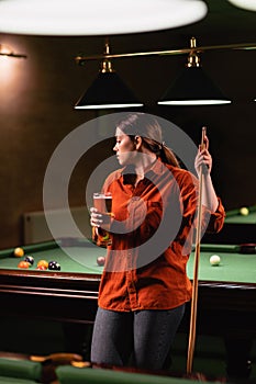 Playing billiards in a bar. Attractive girl with a cue in her hands drinks a glass of beer and thinks about the game