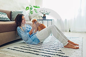 Playing with cat at home. Young woman sitting on carpet and hugging pet