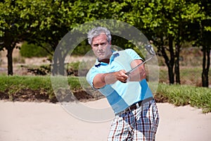 Playing his way out of a ditch. a mature man playing a golf shot from a sand bunker.