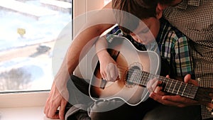 Playing a musical instrument. Dad teaches his son to play the guitar, sitting on the windowsill.