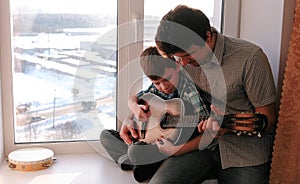 Playing a musical instrument. Dad teaches his son to play the guitar, sitting on the windowsill.