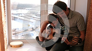 Playing a musical instrument. Dad teaches his son to play the guitar, sitting on the windowsill.