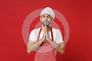 Pleading bearded male chef cook or baker man in striped apron white t-shirt toque chefs hat posing isolated on red