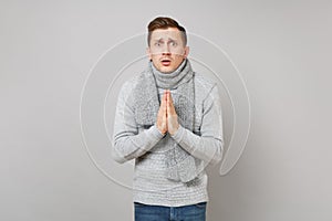 Pleading young man in gray sweater, scarf holding hands together, praying on grey wall background. Healthy