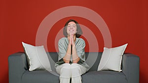 Pleading young woman keep palms together implore beg for favor or help sitting on sofa. Red background