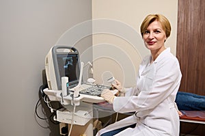 Pleasant adult European woman, radiologist, diagnostics specialist, sitting at ultrasound machine, holding transducer, looking