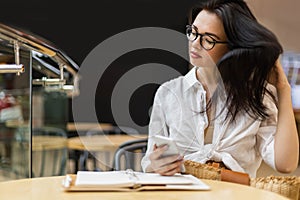 Pleasant Asian business woman chatting use smartphone taking notes in notepad at table in cafe