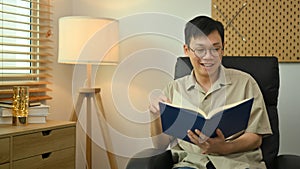 Pleasant asian man wearing glasses and casual clothes reading book on armchair in cozy living room