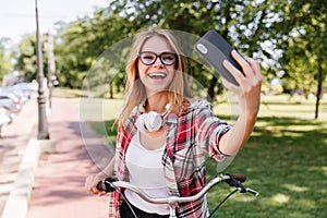 Pleasant blonde girl using smartphone for selfie in park. Charming smiling lady in glasses riding o