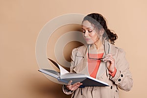 Pleasant brunette woman in casual wear, holding spectacles in her hands and reading a book, on isolated beige background