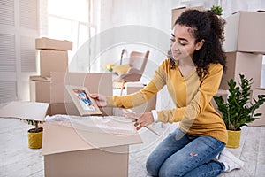 Pleasant curly-haired girl packing framed photo before moving out