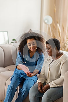 Pleasant curly nurse showing online news to elderly lady