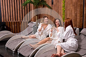 Pleasant ladies enjoy drinking champagne at spa. Three beautiful women wearing bathrobes having cool resting, holidays