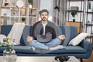 Pleasant likable young arabian indian man, sitting in lotus pose with laptop pc on sofa at living room, and meditating