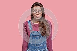 Pleasant looking brunette woman pouts lips, makes grimace at camera, wears purple jumper and denim overalls, wants to