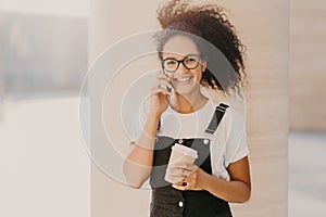 Pleasant looking curly teenage girl wears transparent glasses, talks on mobile phone, has coffee break, dressed in white t shirt