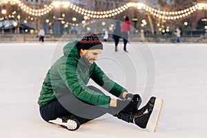 Pleasant looking man wears green coat and hat, sits on ice and l