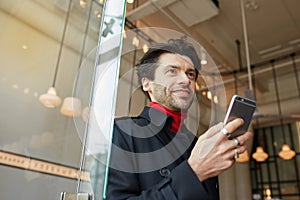 Pleasant looking young attractive dark haired man smiling gladly while keeping smartphone in raised hand, posing over cafe