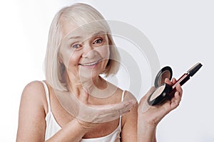 Pleasant retired woman holding face powder and mascara