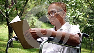 Pleasant senior man sitting in wheelchair and reading a book at summer park.