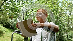 Pleasant senior man sitting in wheelchair and reading a book at summer park.