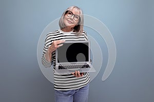 pleasant smiling middle aged 60s woman showing mockup web page on laptop on bright studio background