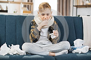 Pleasant tired sick teen boy with scarf around neck, sitting on soft couch and coughing
