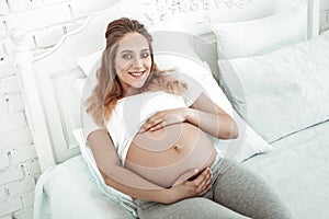 Pleasant woman with wide smile wrapping pregnant belly