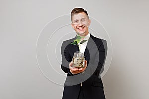 Pleasant young business man in classic black suit hold pile gold coins in glass jar with green plant sprout isolated on