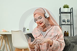 Pleasant young Muslim woman relaxing on couch texting messaging, browsing wireless internet on smartphone