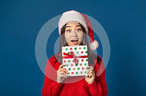 Pleasantly excited happy woman holds in hands christmas gift. Beautiful woman in santa claus hat on blue background