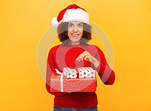 Pleasantly excited happy woman holds in hands christmas gift. woman in santa claus hat on orange background, smiling