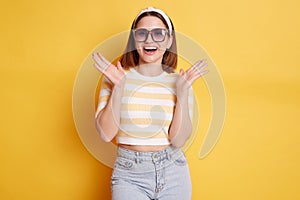 Pleasantly surprised woman wearing striped T-shirt hair band and sunglasses standing isolated over yellow background raised her