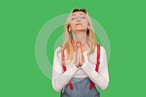 Please, god! Portrait of upset pleading adult woman in denim overalls praying with closed eyes, imploring help