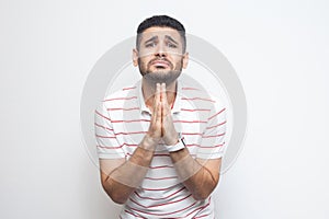 Please help me or forgive. Portrait of sad alone bearded young man in striped t-shirt standing, palm hands and looking at camera