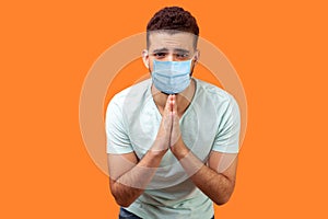 Please, I`m begging! Portrait of upset man with medical mask in white t-shirt holding arms in prayer, asking help or forgiveness