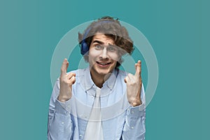 Please. Portrait of a young beautiful man wearing white t-shirt and blue shirt in blue headphones holding his fingers crossed