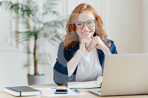 Pleased cheerful red haird female economist develops financial startup project, poses in office interior, works in business sphere