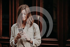 Pleased delighted female youngster watches video on smart phone, reads banking notification, dressed in white raincoat, poses agai