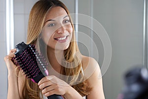 Pleased girl holds round brush hair dryer to style hair in her bathroom at home. Young woman using salon one-step hair dryer and