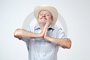 Pleased hopeful mature man closes eyes, keeps palms together, prays to have good luck, has dreamful look