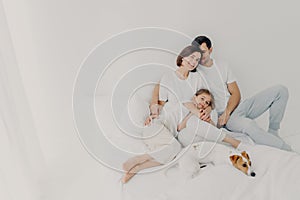 Pleased husband, wife and their small daughter pose in spacious bedroom with favourite pet, have fun, embrace and express love to