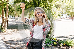 Pleased lady in trendy glasses holding cup of coffee and laughing. Outdoor portrait of stunning eur