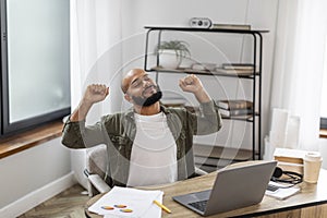 Pleased latin man stretching his back and arms while working on laptop computer, sitting at home office, taking break