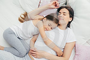 Pleased little child sleeps near her mother, embraces with love, has pleasant dreams, lie on comfortable bed. Mum and cute daughte