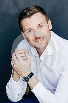 Pleased male entrepreneur, bristle, dark hair, hands together, white shirt, confident expression. Isolated on dark wall